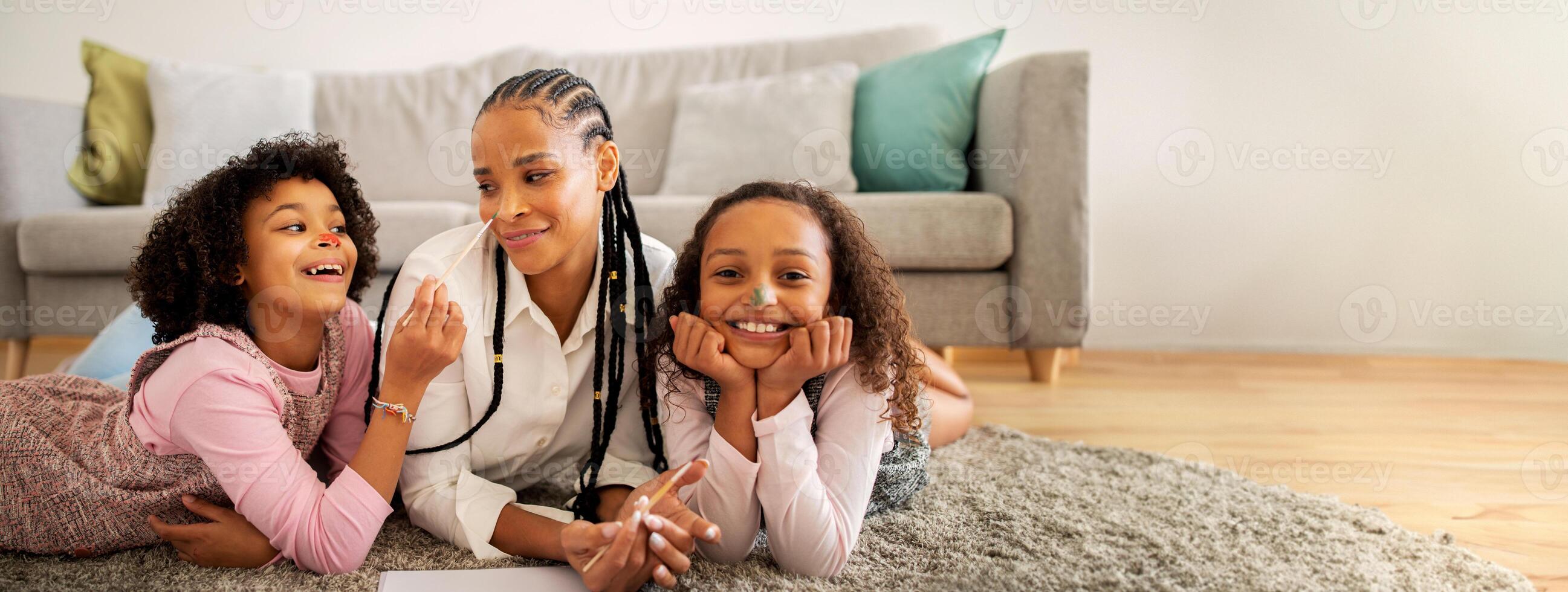 A black mother interacts joyfully with her two daughters, who are playfully lying on the floor in their stylish living room, filled with warmth and family bonding. photo