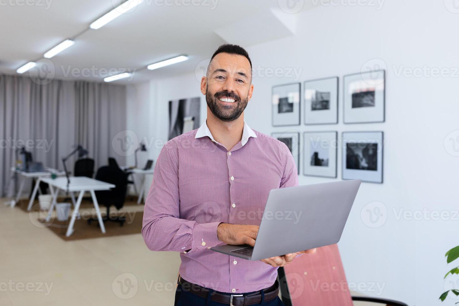 A cheerful man stands in a contemporary office, holding a laptop and engaging with his work. The well-lit space features stylish decor and organized workstations in the background. photo