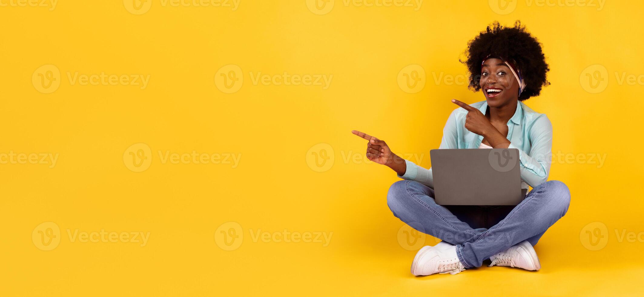 Cheerful Black Woman With Laptop Pointing Fingers Aside At Empty Space For Advertisement Text Posing Sitting On Floor Over Pink Background. Studio Shot. Look There Concept photo
