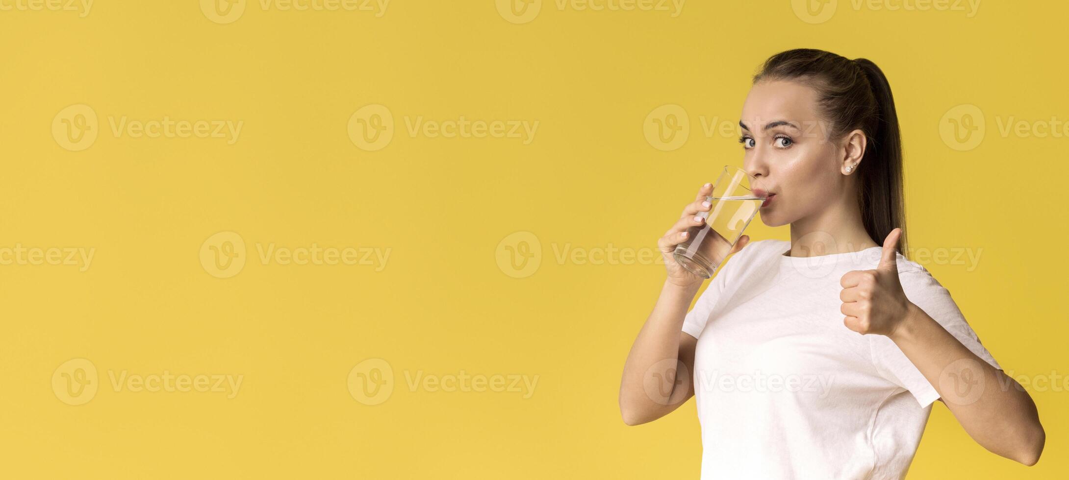 Healthy Liquid. Beautiful Young Woman Drinking Mineral Water And Showing Thumb Up Gesture, Standing On Yellow Background With Empty Space photo