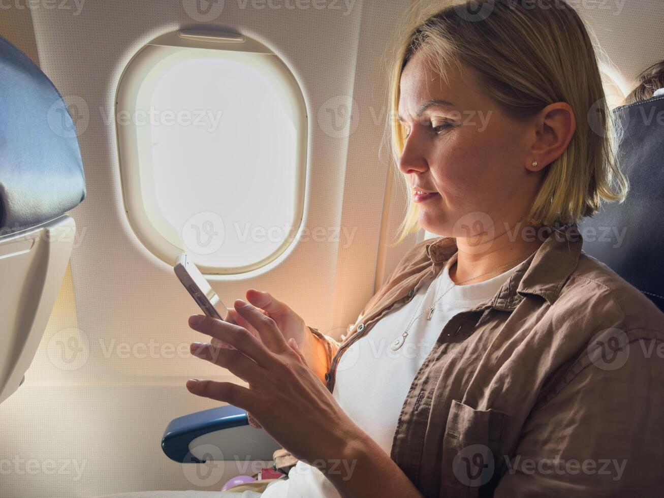Passenger woman is flying in plane. Girl using phone sitting by airplane window. Traveling female with smartphone inside plane enjoying flight. Woman using wifi connection on board. Traveling girl photo
