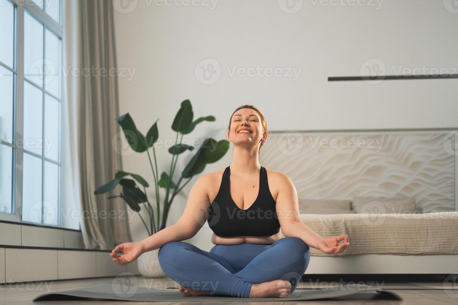 Yoga mindfulness meditation. Woman practicing yoga in living room at home. Plus size female sitting in lotus pose meditating smiling relaxing indoor. Girl doing breathing practice. Yoga at home photo