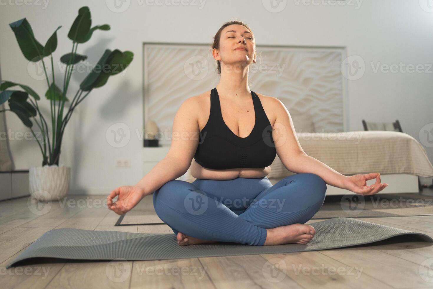Yoga mindfulness meditation. Woman practicing yoga in living room at home. Plus size female sitting in lotus pose meditating smiling relaxing indoor. Girl doing breathing practice. Yoga at home photo