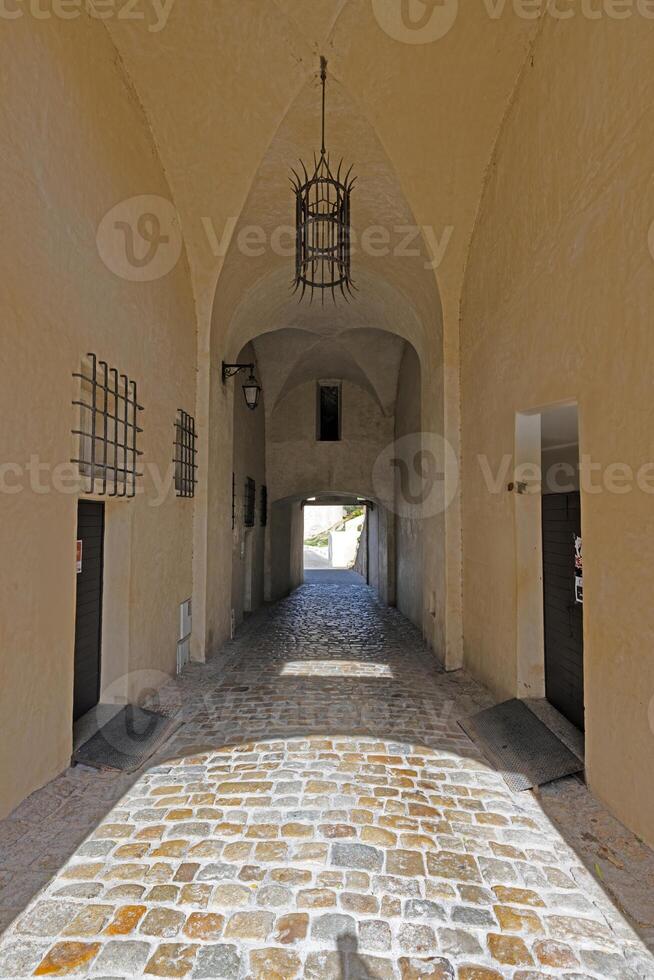 An arched stone corridor with a cobblestone floor, dim lighting, and metal lanterns hanging from the ceiling photo