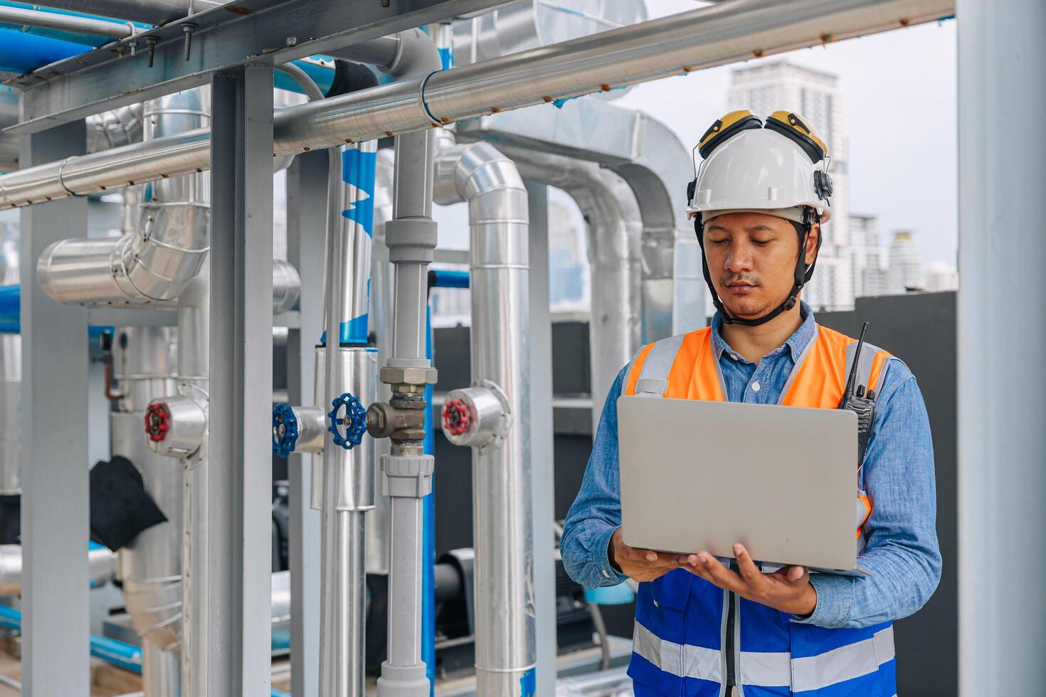 engineer man in a safety vest is standing around gas plump pipe, looking at a laptop. He is wearing a hard hat and a helmet photo
