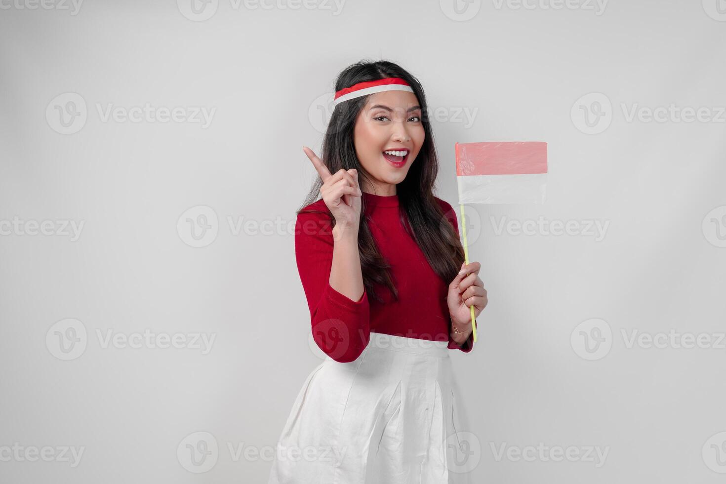 Cheerful young Asian woman is holding mini country flag and pointing to the copy space upwards. photo