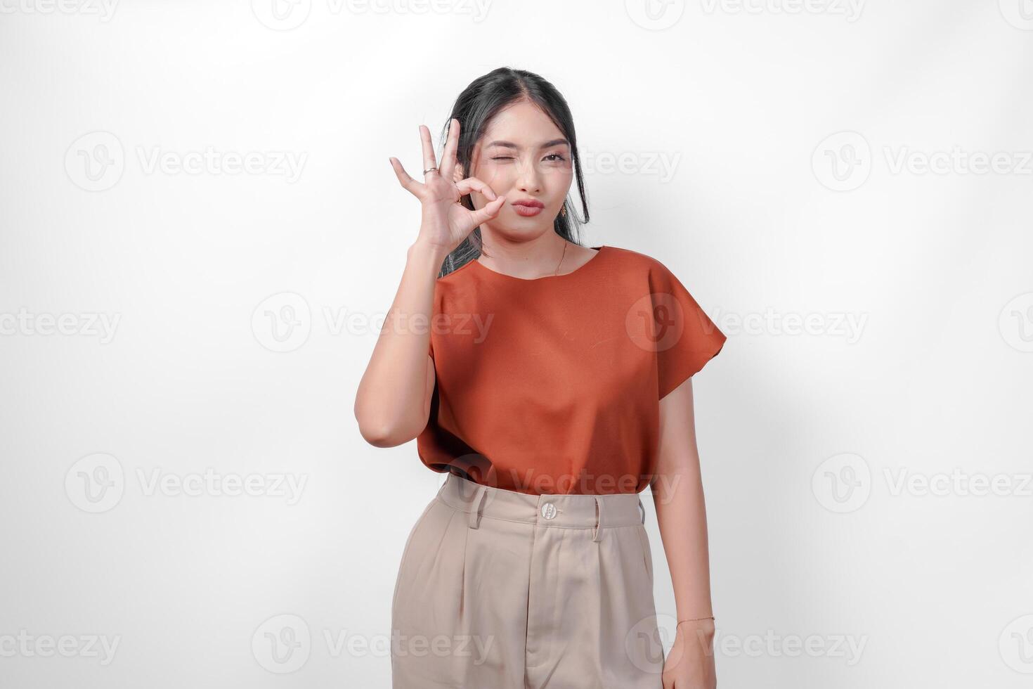 A happy young Asian woman in a brown shirt and taupe pants giving okay good hand gesture, isolated by white background. photo