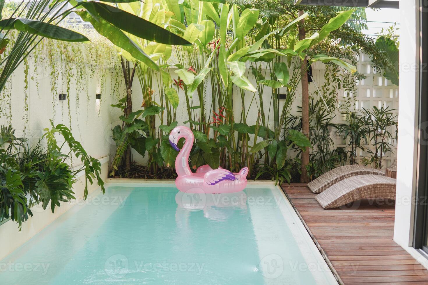 A pink inflatable flamingo floats in a pool. The pool is in a backyard, and there are two lounge chairs on the deck. photo