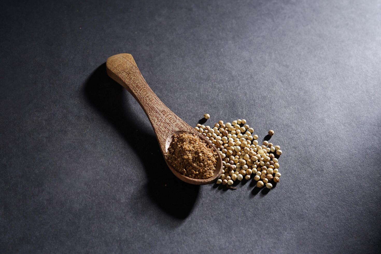 coriander powder in wooden spoon isolated on dark background, placed over a heap of whole coriander seeds, spice concept photo