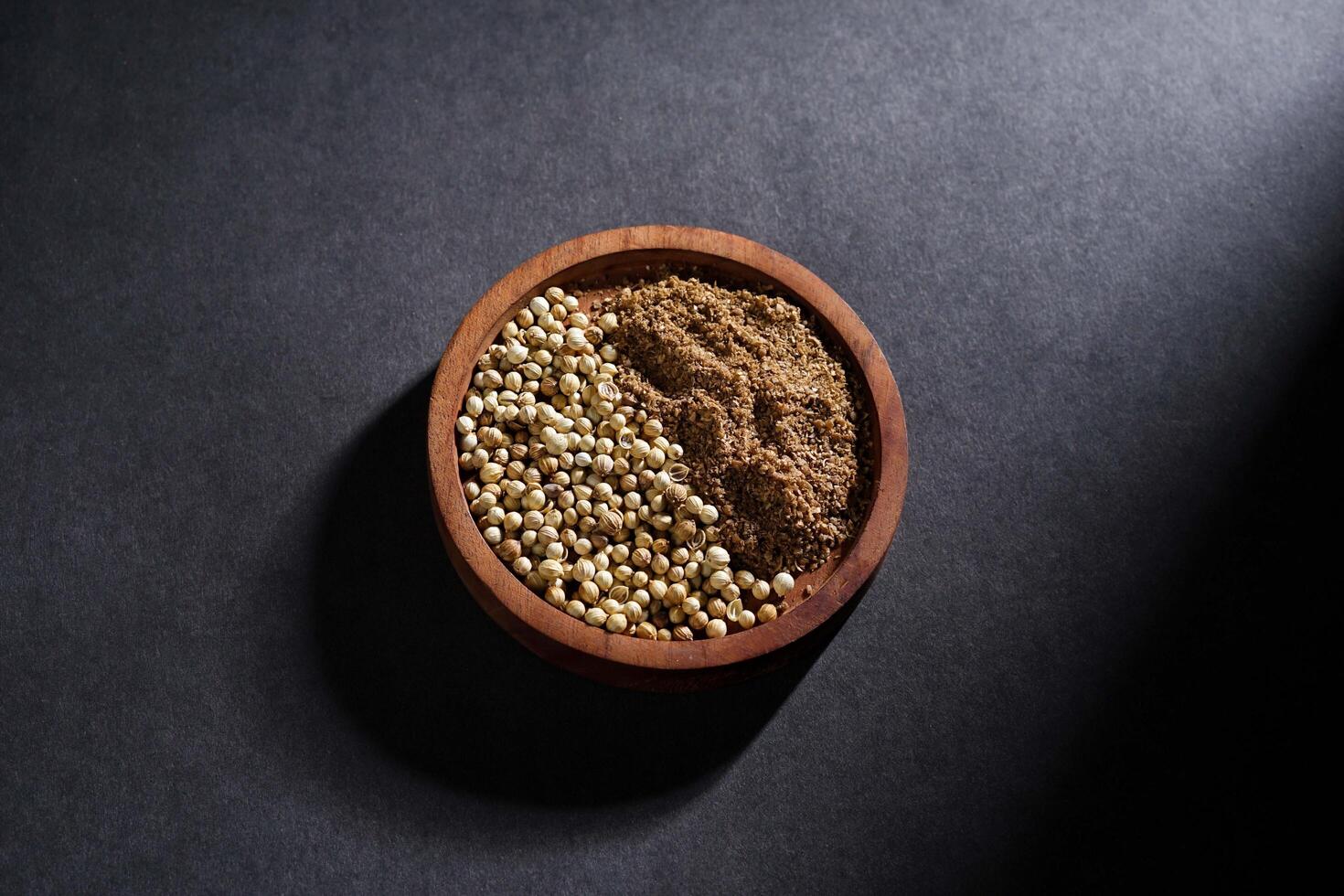 coriander seed and coriander powder in wooden bowl isolated on dark background, spice concept photo