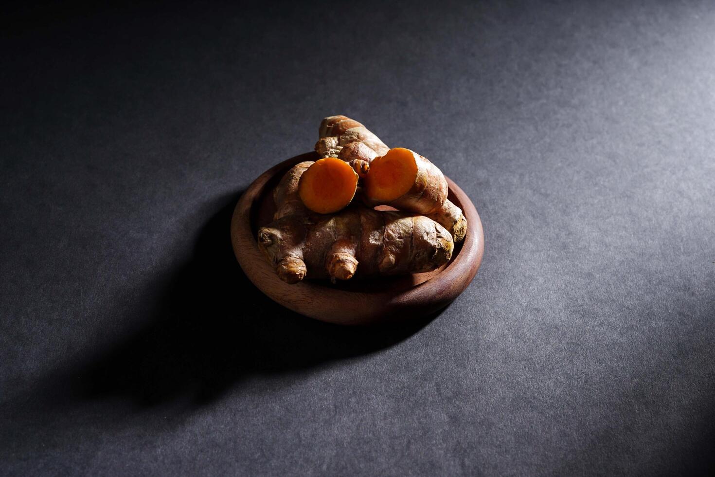 Fresh turmeric root and slices in wooden bowl isolated on dark background, turmeric Curcuma longa Linn, spice concept photo