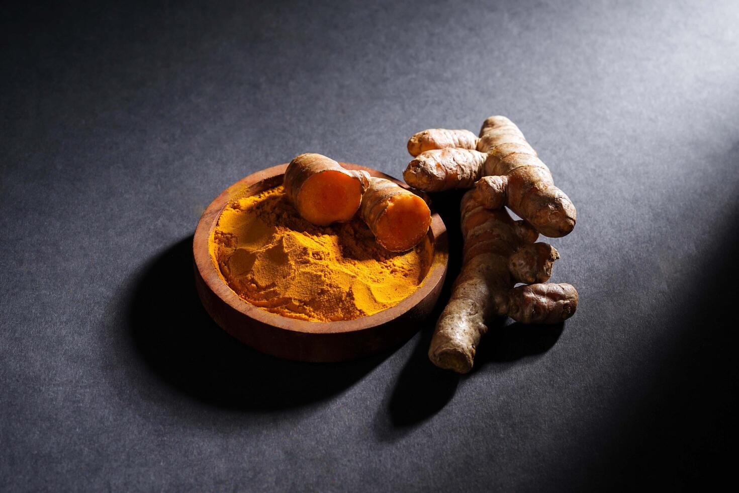 Fresh turmeric root and turmeric powder in wooden bowl, turmeric isolated on dark background, spice concept photo