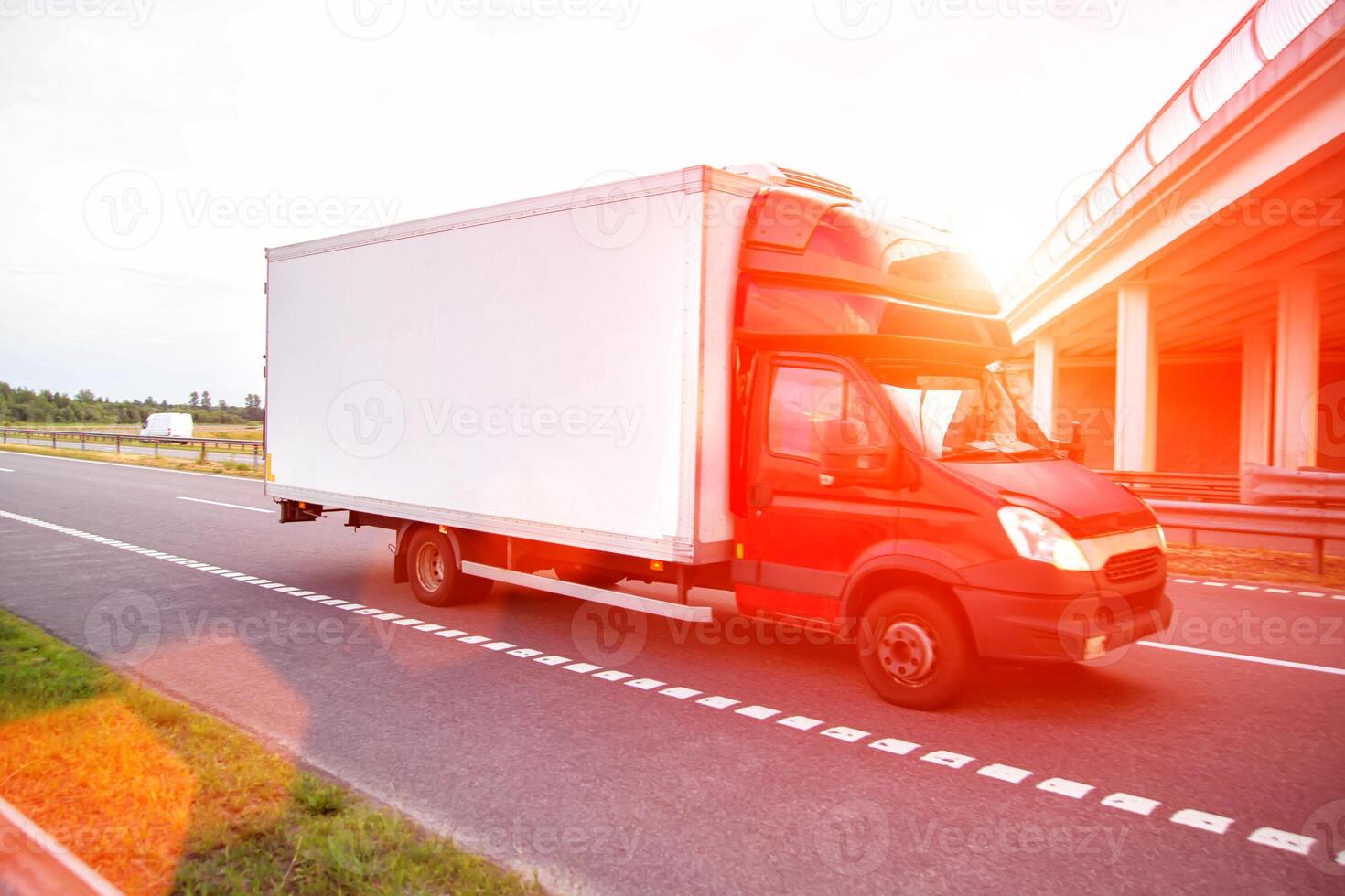 A red light-duty refrigerated van transports perishable cargo on the highway in the summer against the backdrop of sunset, industry photo