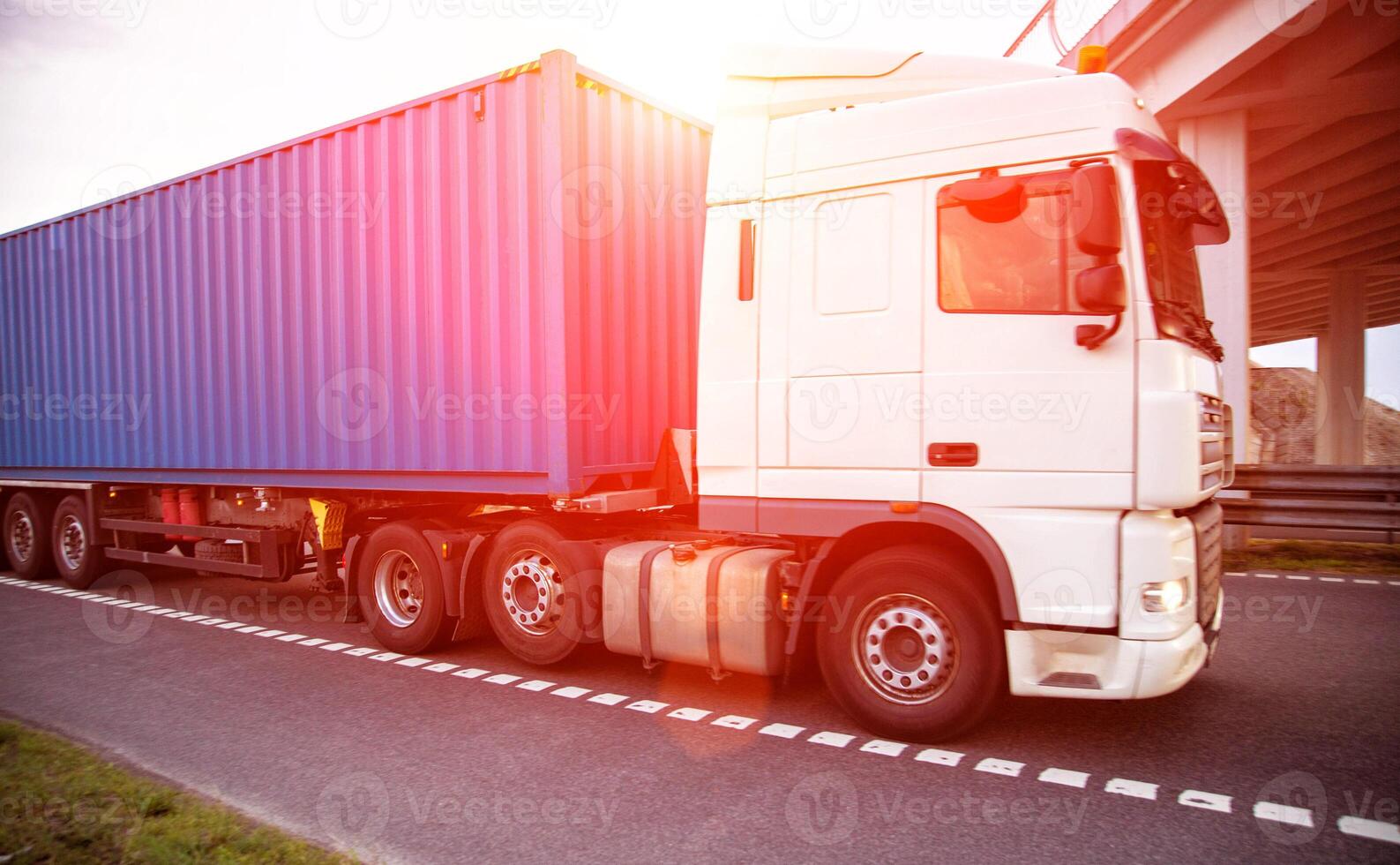 A container truck with a semi-trailer transports cargo from a port to another country along a highway in the summer against the backdrop of the sun. Logistics in cargo transportation photo