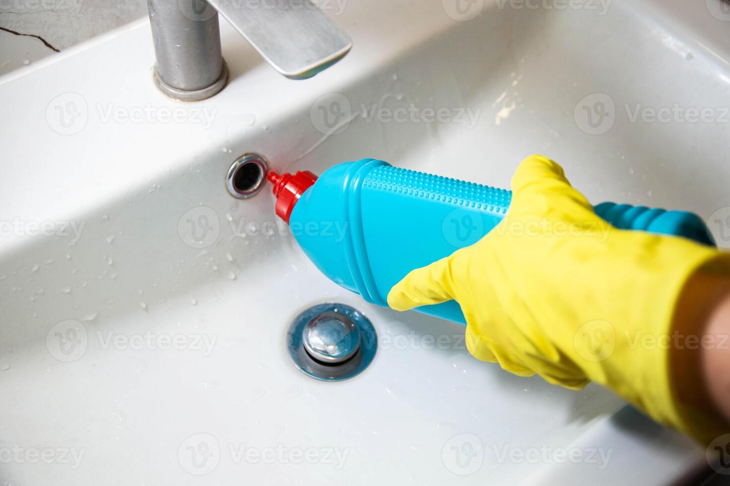 The hands of a girl in a yellow cap with a blue bottle of detergent clean and wash the sink from limescale and dirt. Cleaning company services, sanitary equipment washing. Copy space for text photo