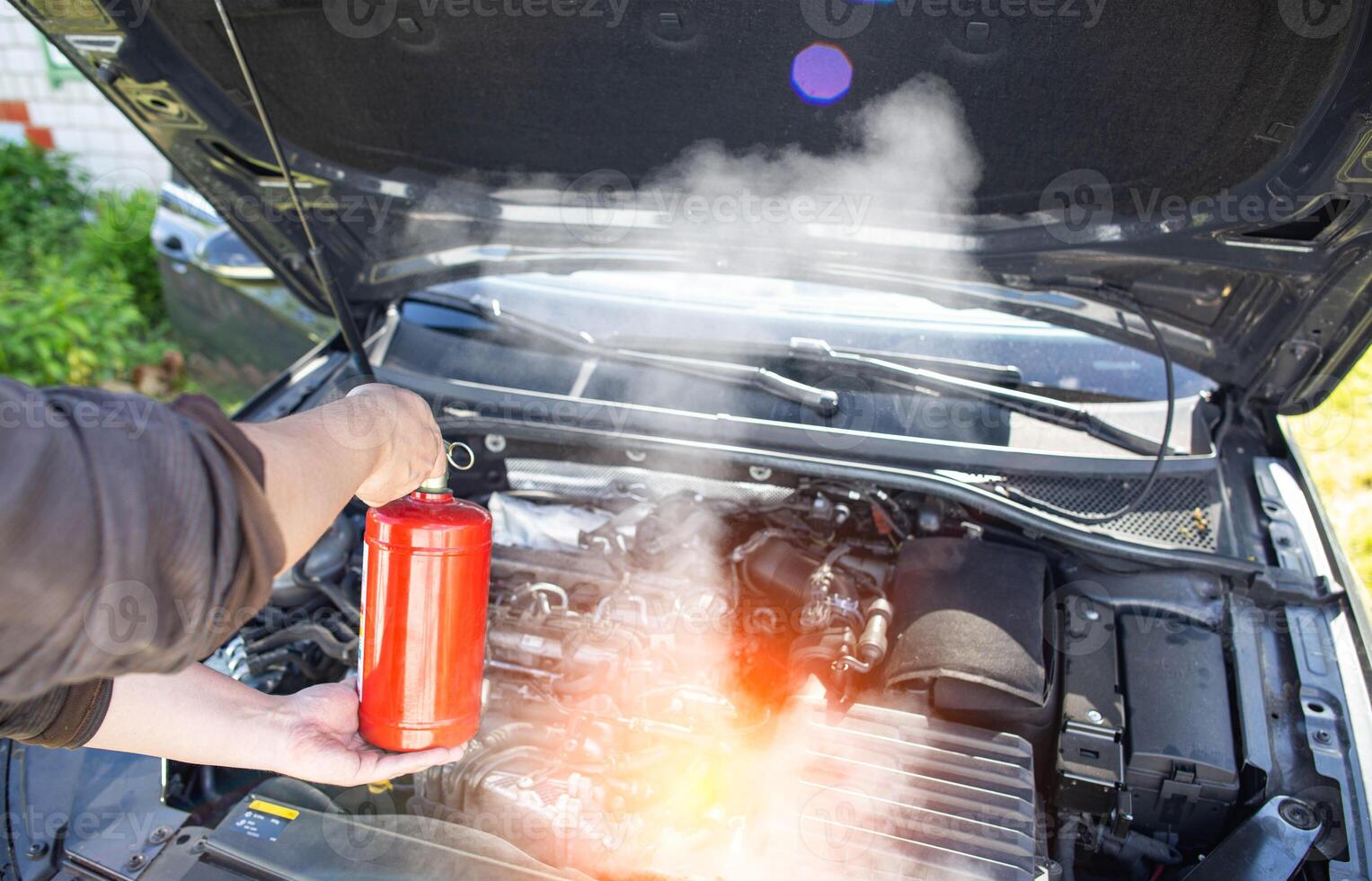 The driver of the car directs a powder fire extinguisher at the engine compartment of the car in which there is fire and smoke. Car fire, background. Electrical short circuit photo