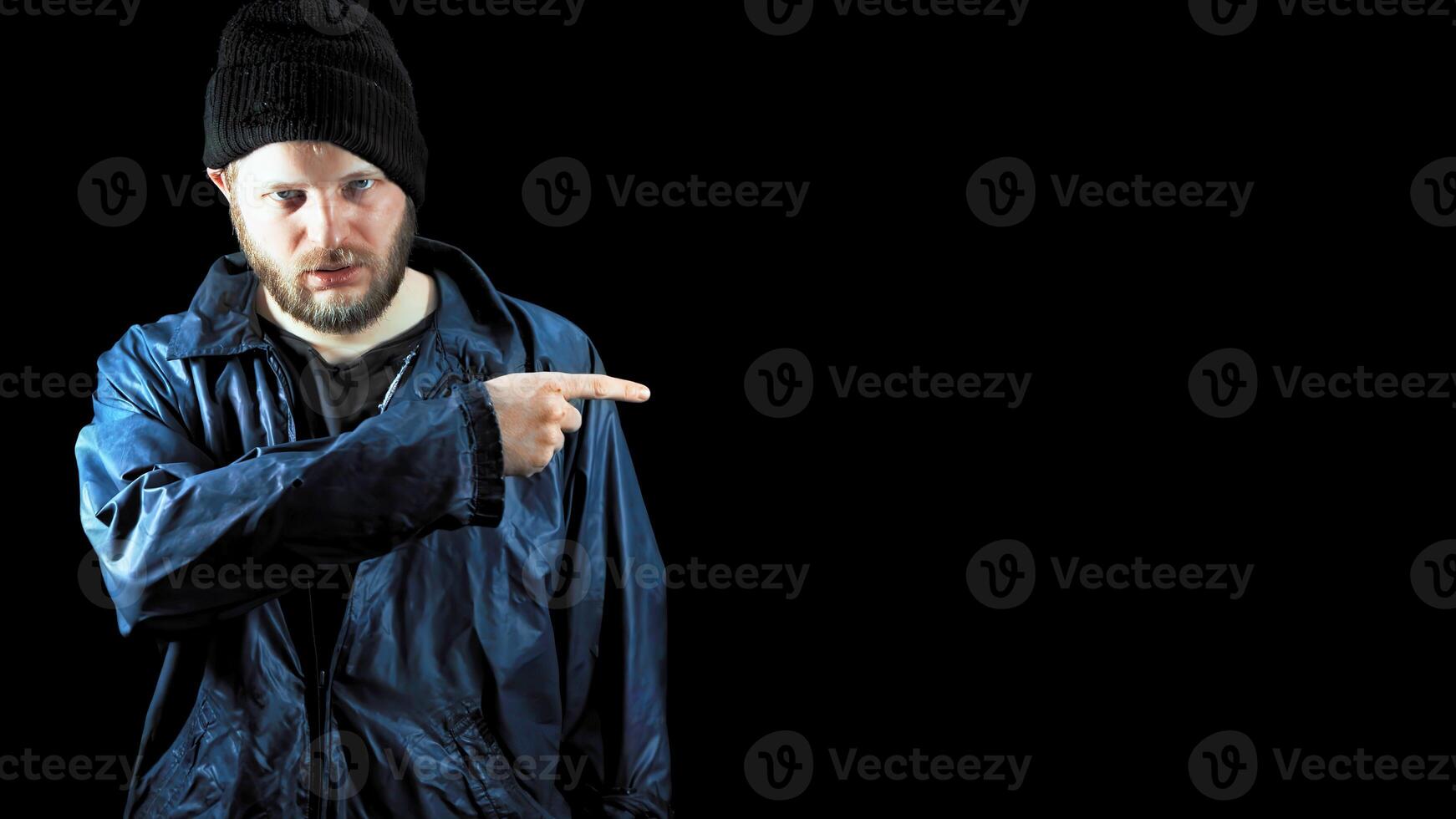 Bearded homeless points to empty space on black background, copy space. Tramp in dirty clothes. Male refugee. Drunkenness, alcoholism. Below poverty line concept. Fugitive beggar. Illegal immigrant photo