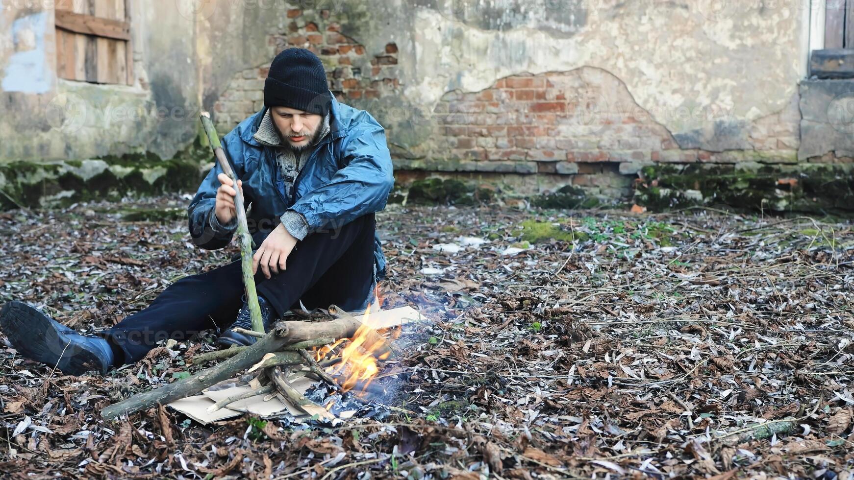 Homeless man sits on ground and uses stick to keep fire burning to keep warm from cold. Tramp in dirty clothes and hat cap. Below poverty line. Illegal immigrant. Male refugee. Fugitive beggar photo