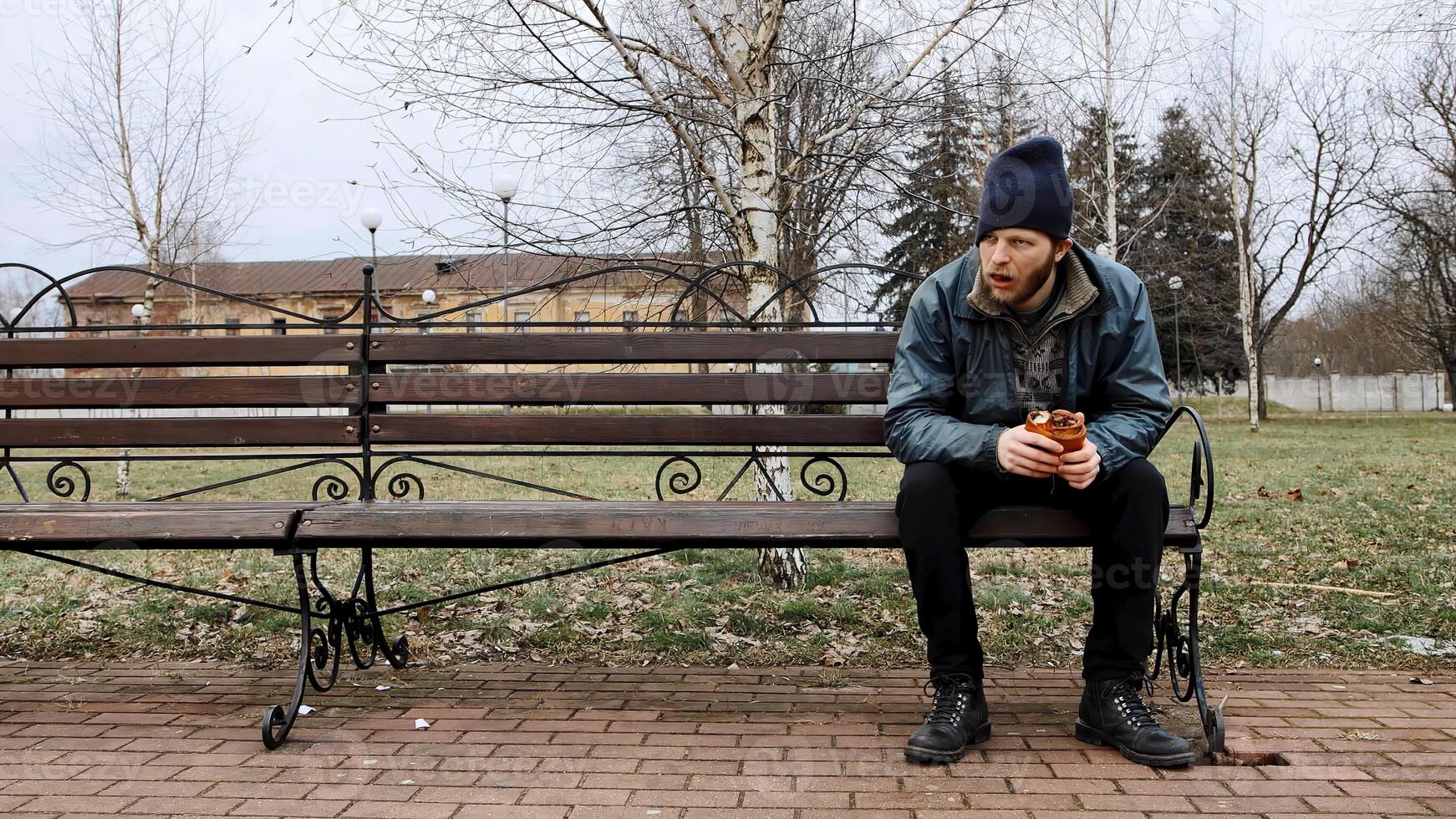 Hungry bearded homeless man sits on a bench and eats a bun in a city park. Below poverty line. Male tramp in dirty clothes with cap and hat. Beggar. Illegal immigrant. Drunkenness, alcoholism photo