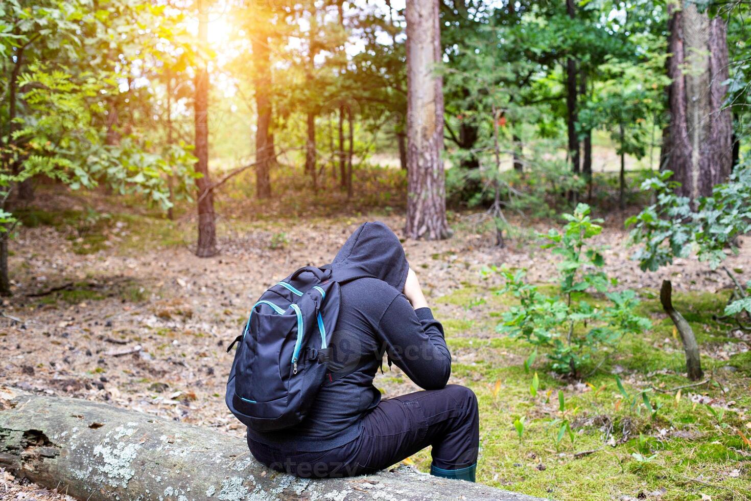 A man with a backpack in a hood sits on a fallen tree in the middle of a clearing in the forest. Survival in the wild. Dehydration of the body. Mushroom picker lost in the forest. photo