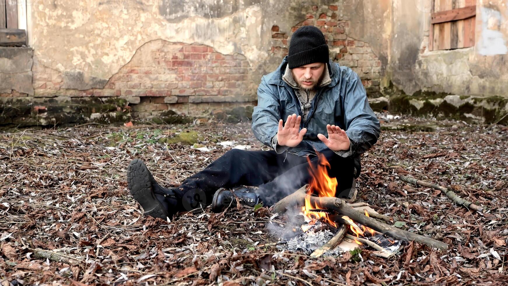 Homeless man sits on ground and pushes pieces of cardboard into fire to keep warm from cold. Tramp in dirty clothes and hat cap. Below poverty line. Illegal immigrant. Male refugee. Fugitive beggar photo