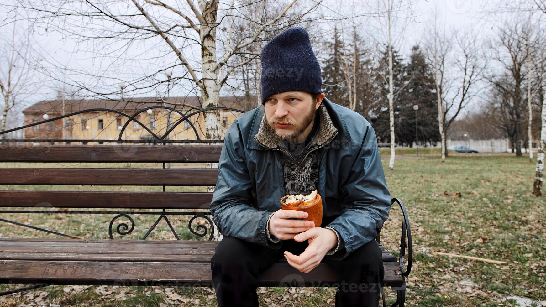 Hungry bearded homeless man sits on a bench and eats a bun in a city park. Below poverty line. Male tramp in dirty clothes with cap and hat. Beggar. Illegal immigrant. Drunkenness, alcoholism photo