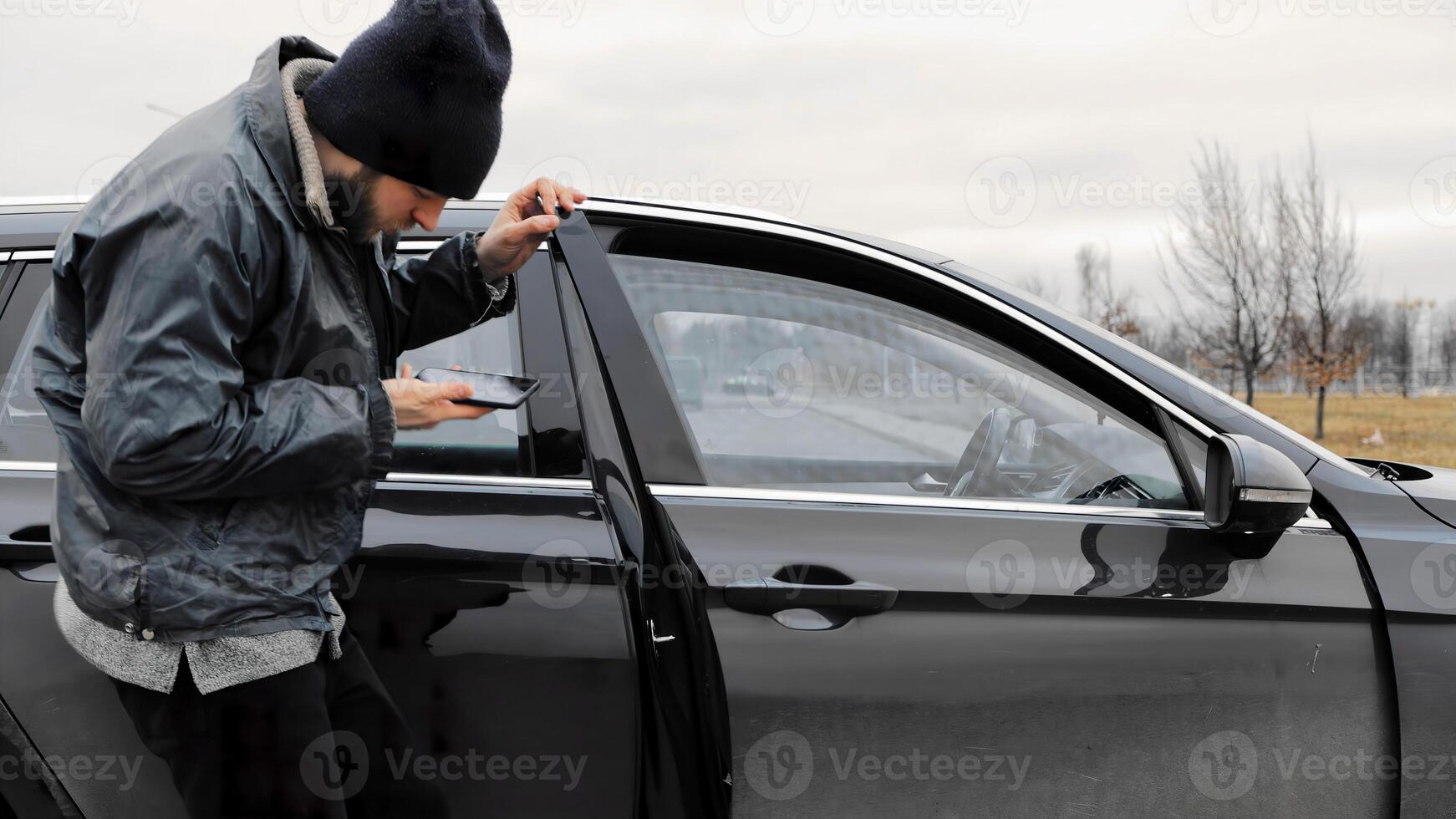 Homeless thief steals smartphone from car in public parking. Auto with unlocked doors open. Property insurance against unlawful theft or crime. Male burglar looks through crack in window photo