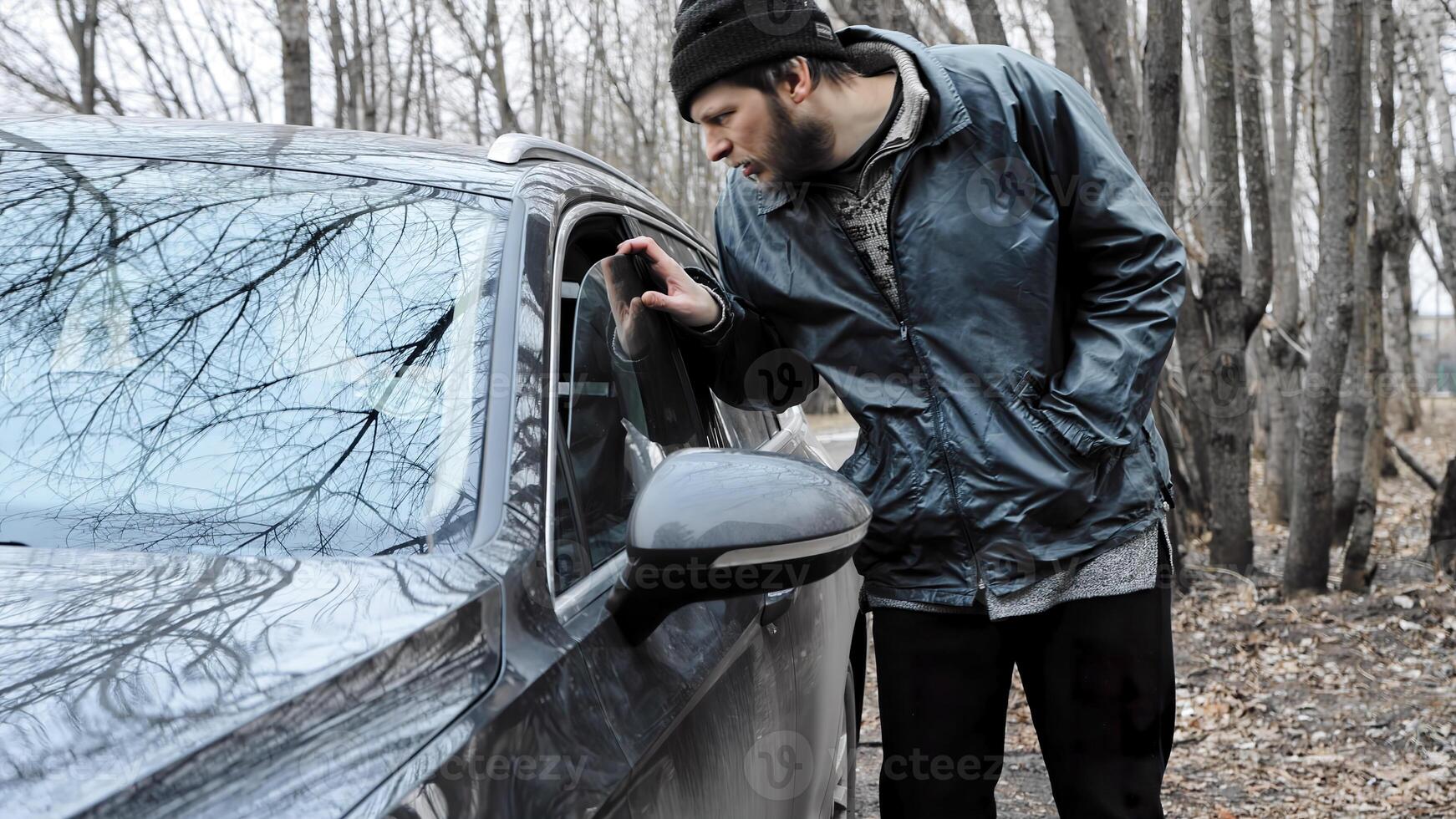 Homeless thief steals purse valuable items from car with unlocked doors open. Property insurance against unlawful theft or crime. Male burglar looks through crack in window. Auto without alarm system photo