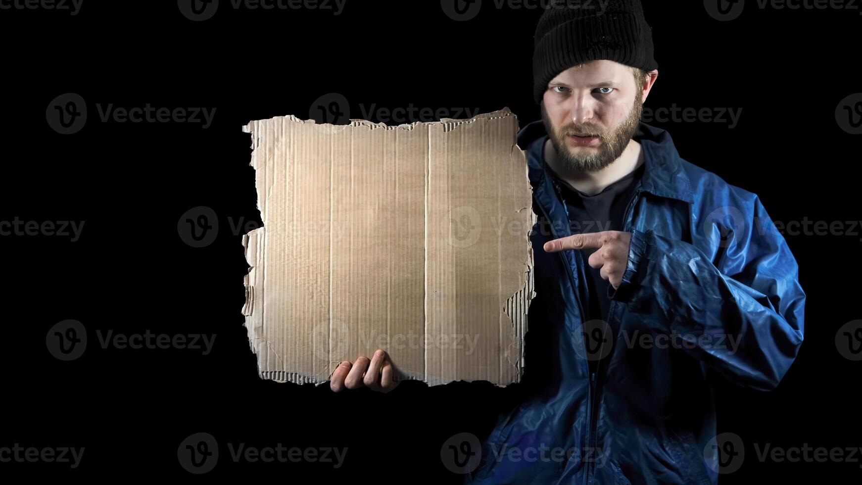Homeless man holds piece of empty clear cardboard. Male tramp in dirty clothes on black background. Poverty. Immigrant. Drunkenness. Refugee is in dark. Bearded fugitive beggar photo