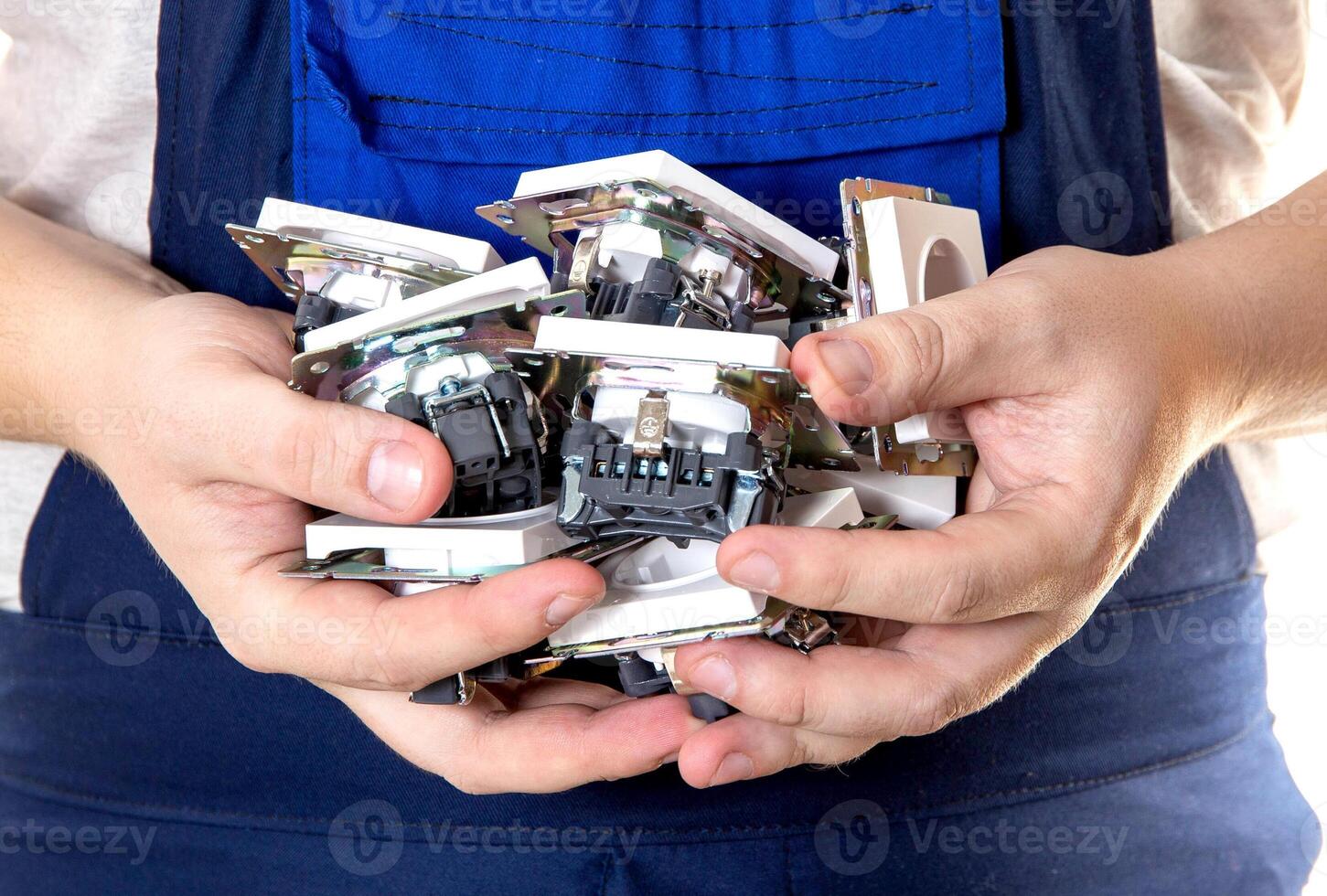 The master holds a lot of electrical sockets and switches in his palms against the background of a blue overalls. Installation and transfer of switches and sockets. Price for electrical work, close-up photo