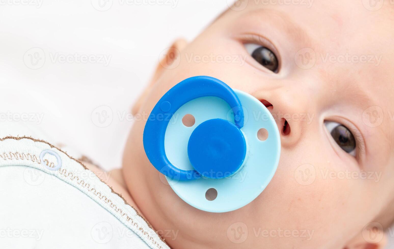 A 5 month old baby with a blue pacifier in his mouth looks at the camera. The concept of the habit of sucking on the nipple, the sucking reflex in babies. Copy space for text photo