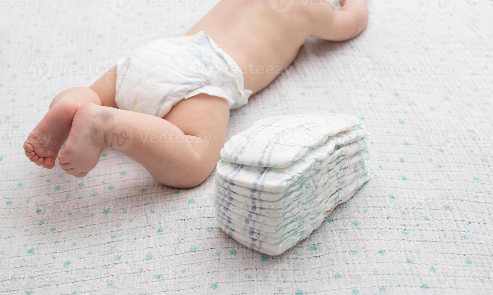 The legs of a baby in a diaper against the background of a stack of children's comfortable diapers, close-up. Copy space for text photo