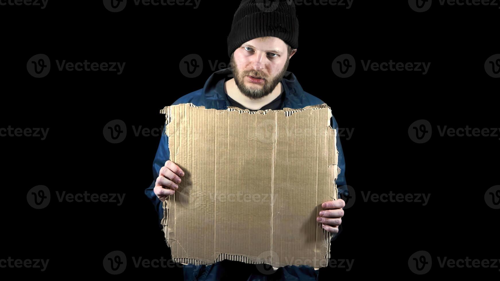 Homeless holds piece of empty clear cardboard. Refugee stands in dark. Male tramp in dirty clothes on black background. Poverty. Bearded fugitive beggar. Immigrant. Drunkenness photo