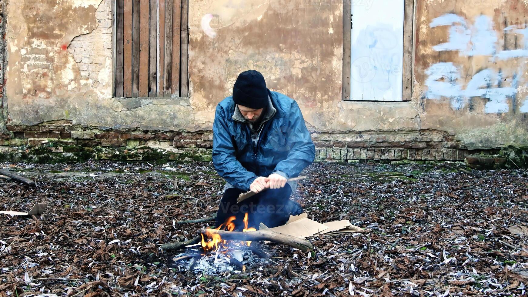 Homeless man sits on ground and pushes pieces of cardboard into fire to keep warm from cold. Tramp in dirty clothes and hat cap. Below poverty line. Illegal immigrant. Male refugee. Fugitive beggar photo