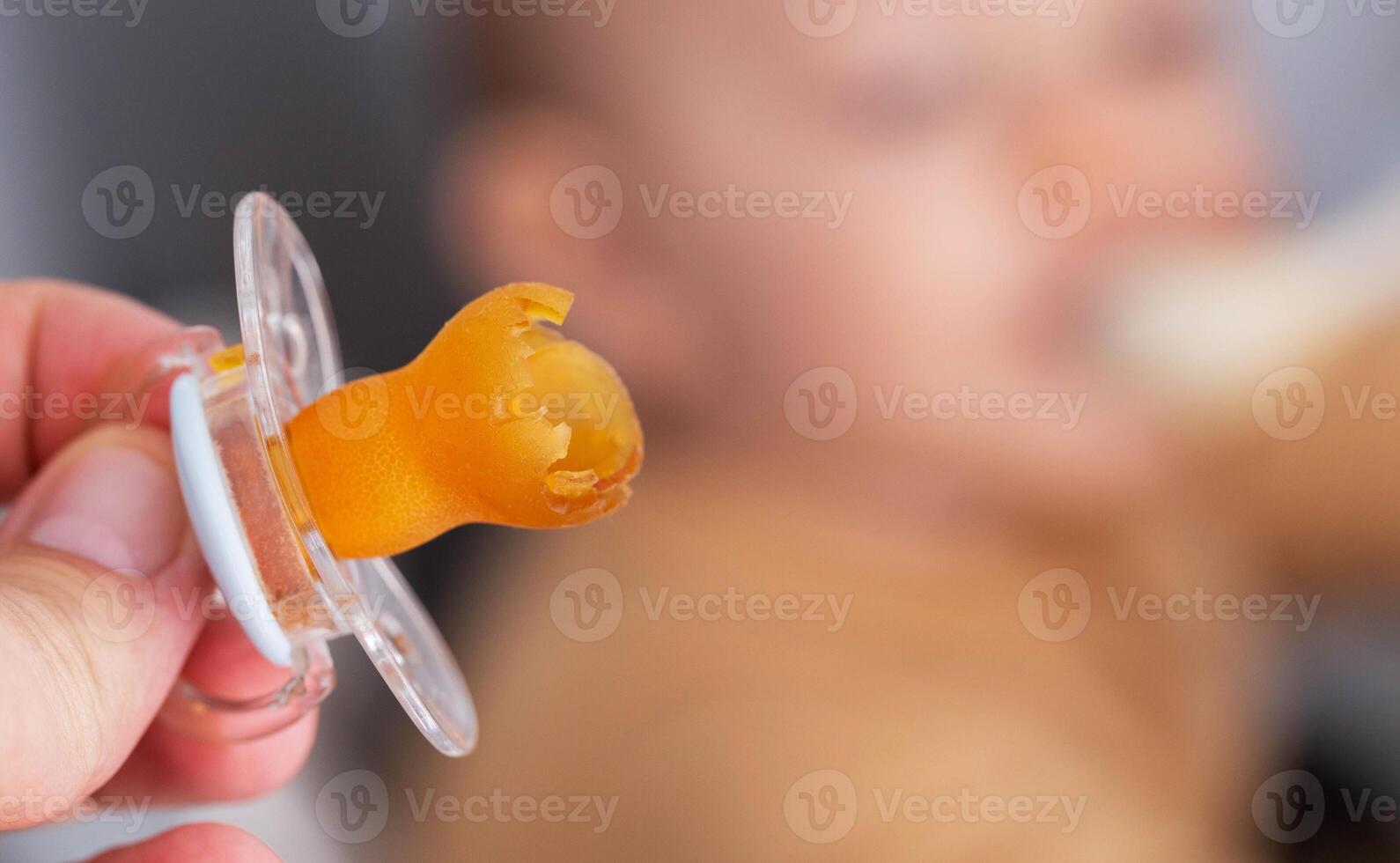 A torn and chewed silicone nipple in the hand against the background of the baby s face. Concept of teething in babies. Sucking reflex, weaning from the pacifier. Copy space for text photo