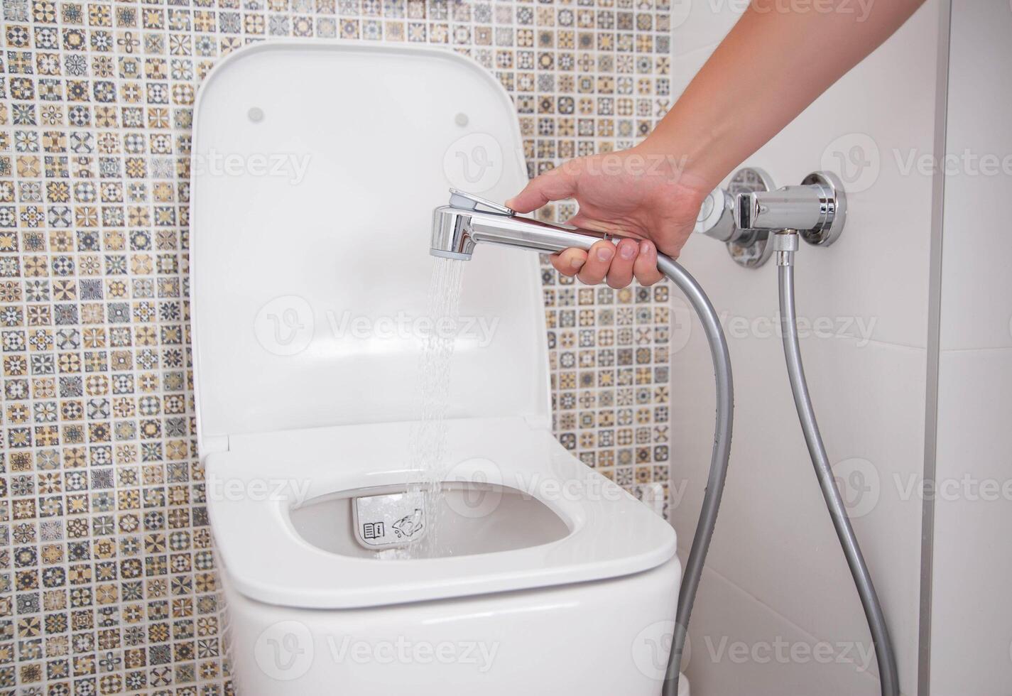 A man holds a hygienic shower against the background of the toilet bowl. Intimate body care and hygiene concept, close-up photo