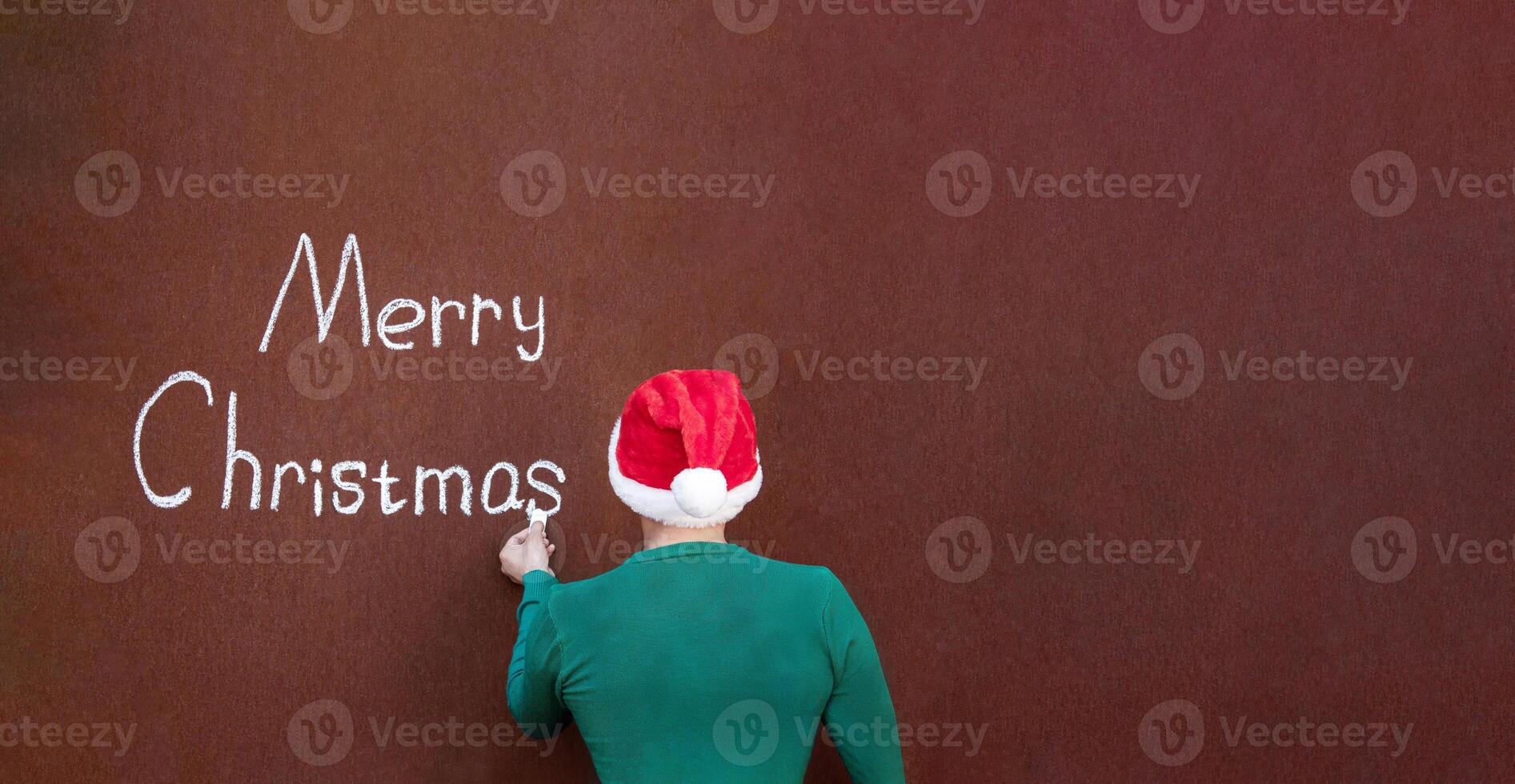 A man writes Merry Christmas with chalk on an abstract surface wearing a Santa hat, Merry Christmas written in chalk on an abstract surface photo