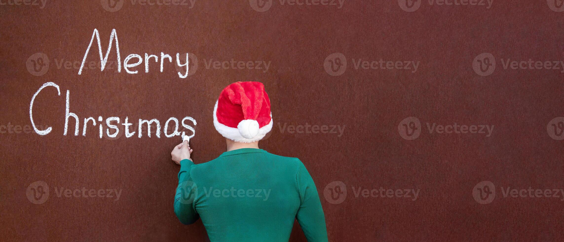 A man writes Merry Christmas with chalk on an abstract surface wearing a Santa hat, Merry Christmas written in chalk on an abstract surface photo