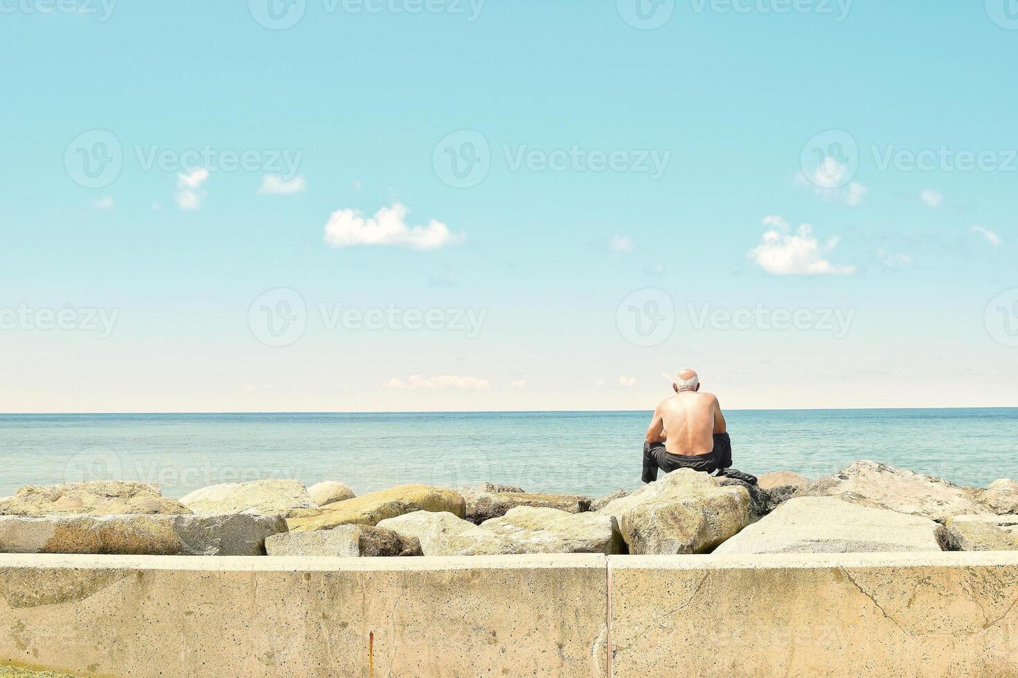 Old man and the sea. An old man sitting on big stone on the seaside. photo