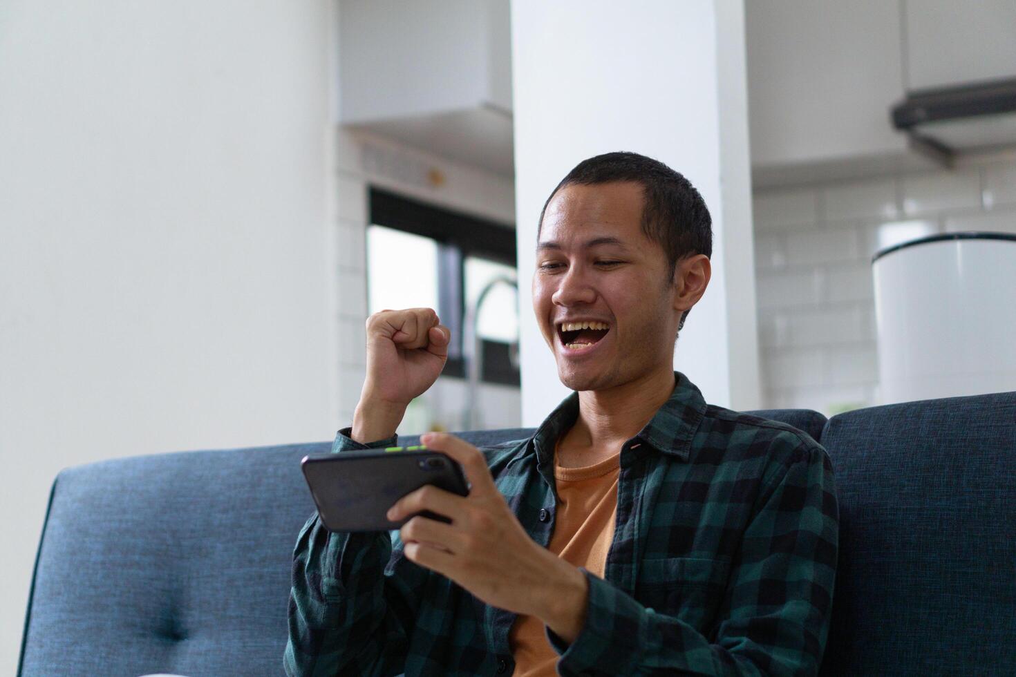Young asian men wearing orange shirt and flannel make fist gesture while playing mobile game on sofa photo