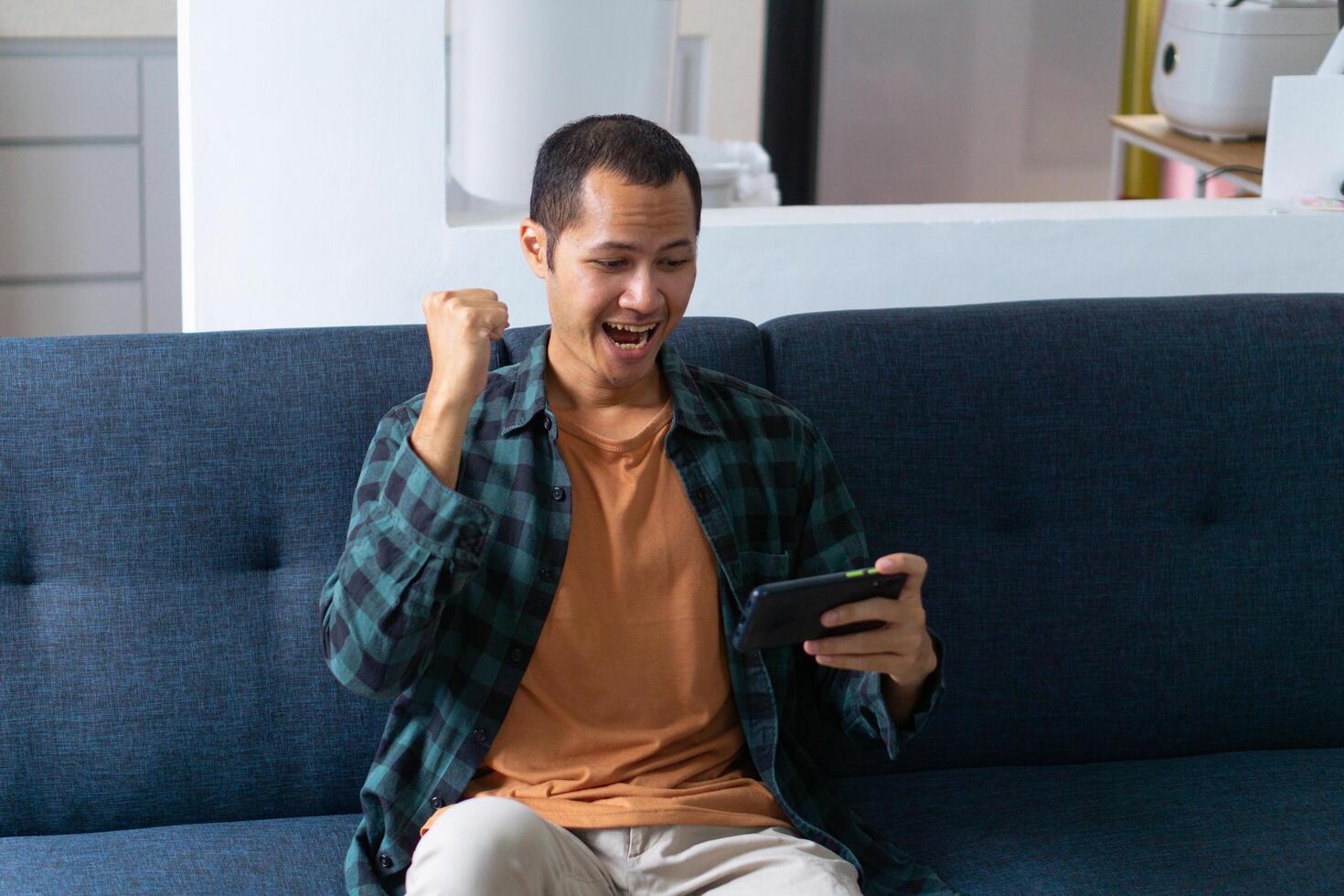 Young asian men wearing orange shirt and flannel make fist gesture while playing mobile game on sofa photo