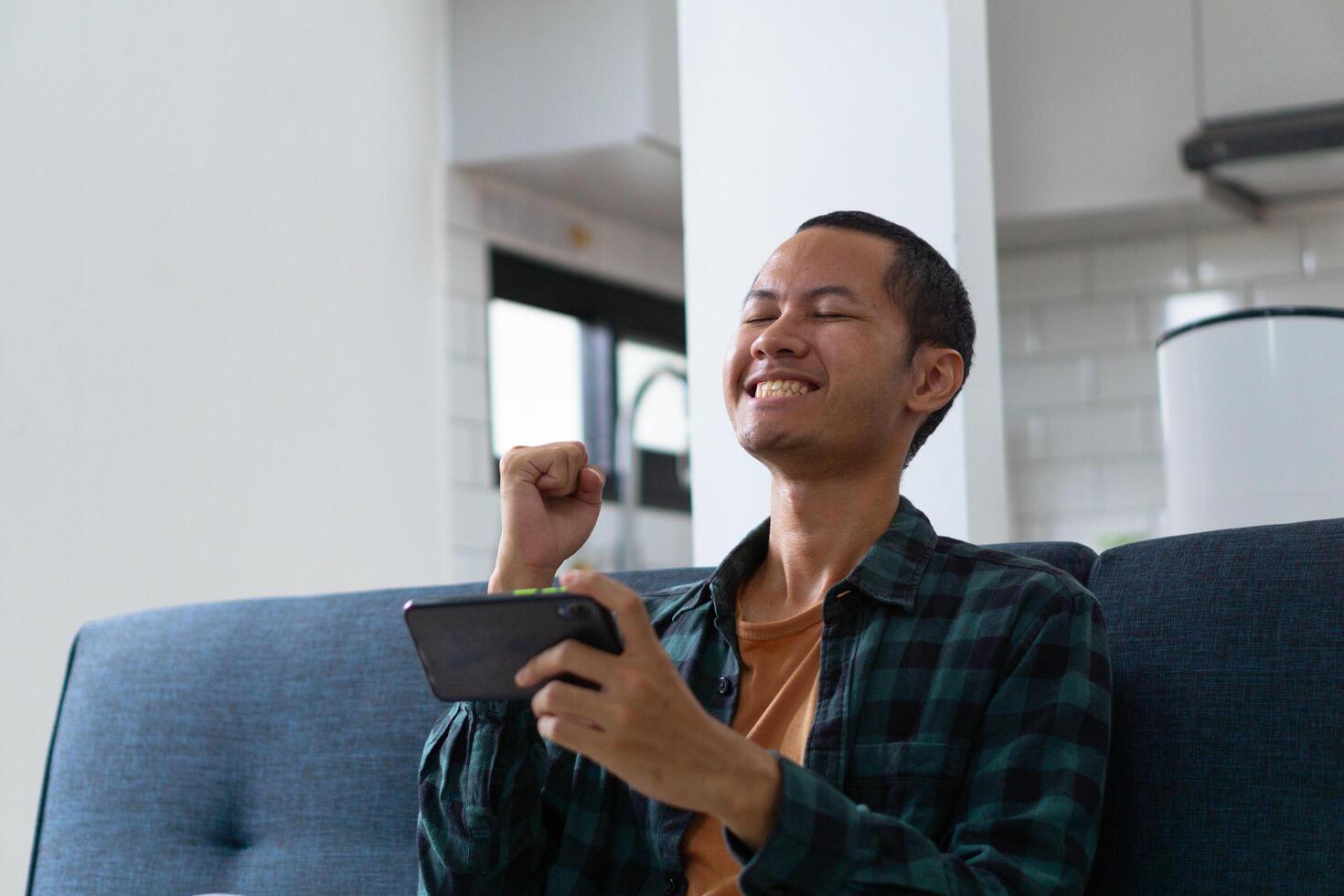 Young asian men wearing orange shirt and flannel make fist gesture while playing mobile game on sofa photo