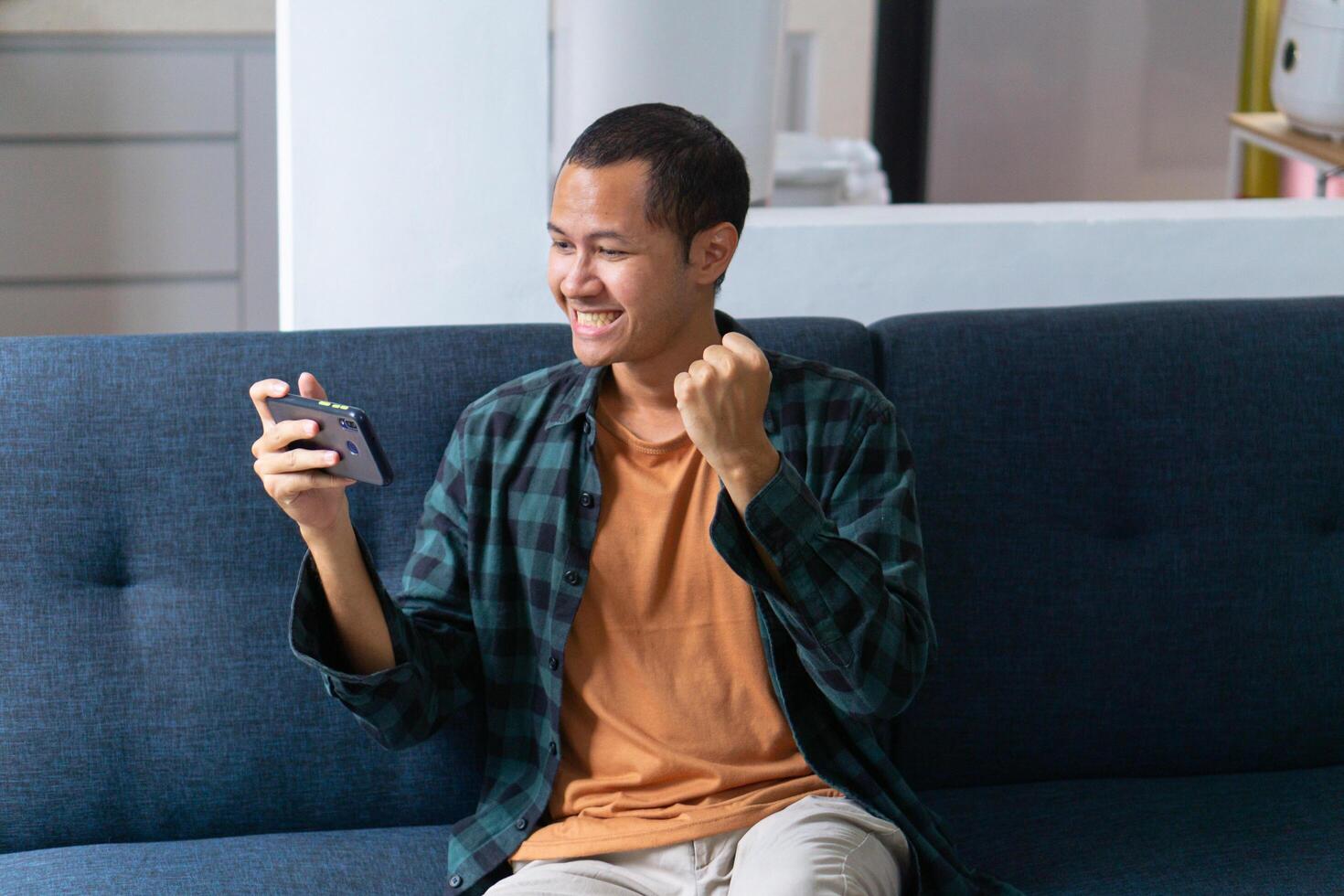 Young asian men wearing orange shirt and flannel make fist gesture while playing mobile game on sofa photo