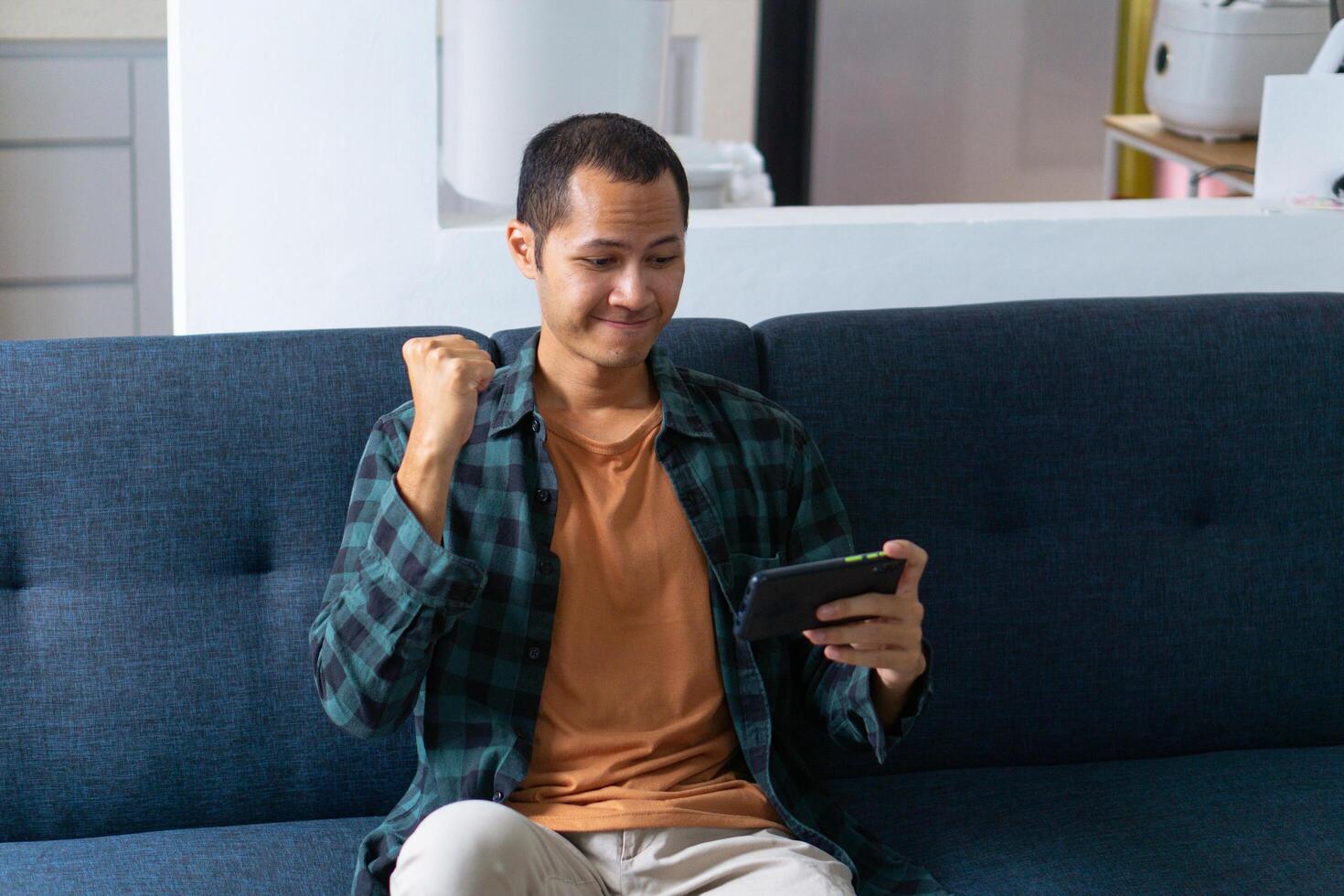 Young asian men wearing orange shirt and flannel make fist gesture while playing mobile game on sofa photo