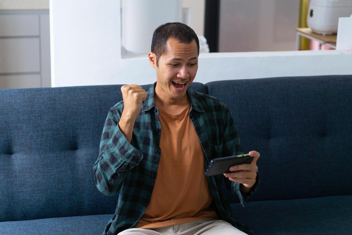 Young asian men wearing orange shirt and flannel make fist gesture while playing mobile game on sofa photo