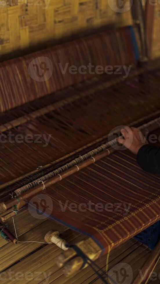 A Baduy woman weaves in front of her home using traditional tools. Her skilled hands create beautiful patterns, preserving cultural heritage and artistry. photo