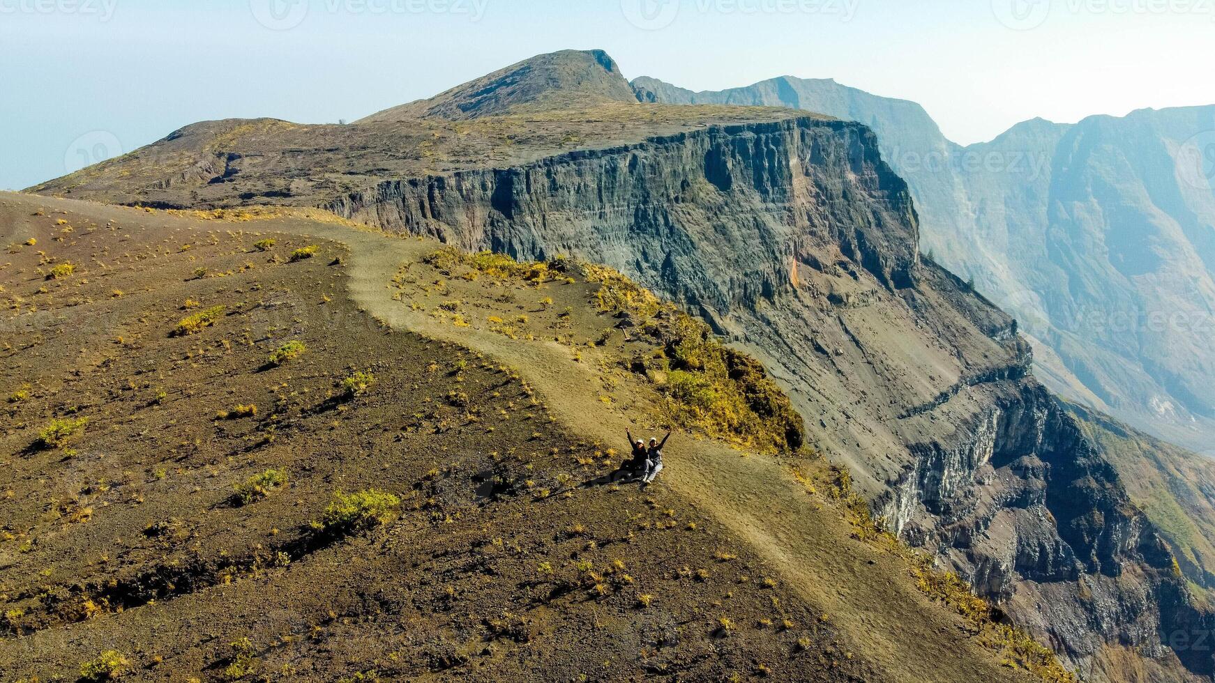An aerial view of Mount Tambora's peak reveals a breathtaking landscape under a blue sky, as the sun rises, casting golden hues over the stunning terrain. photo