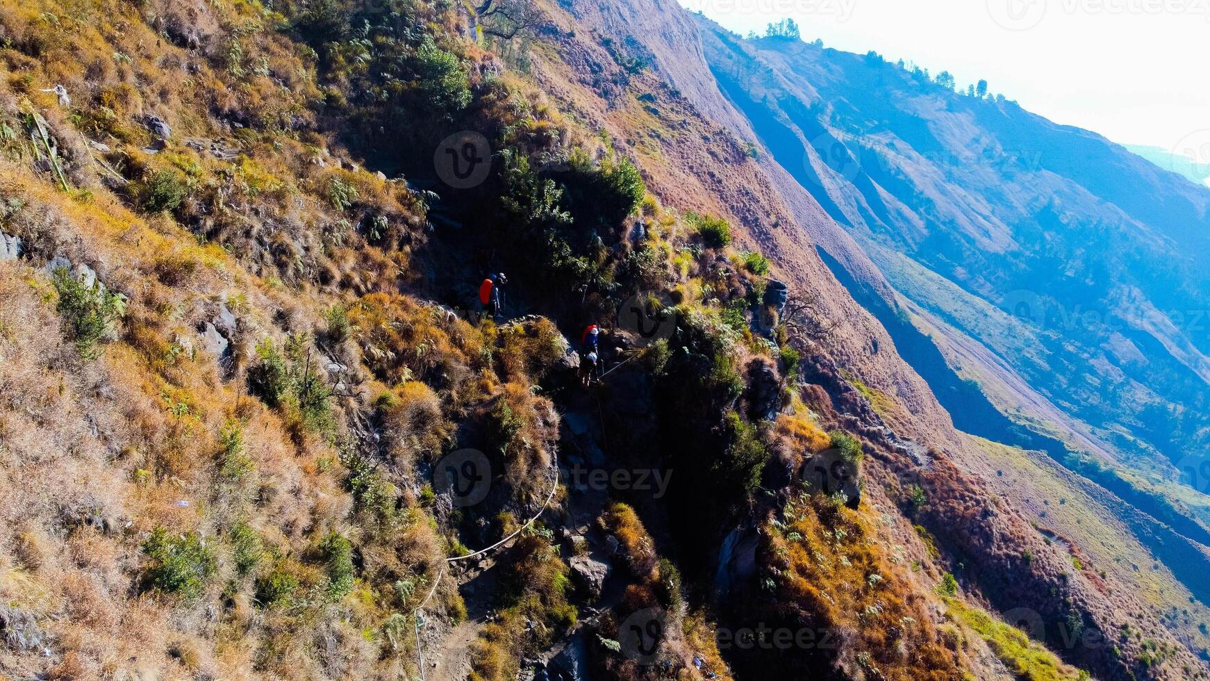 On the trek to Mount Rinjani's summit, a path of lush green and brown grass unfolds beneath a brilliant blue sky. Rest stops dot the route. photo