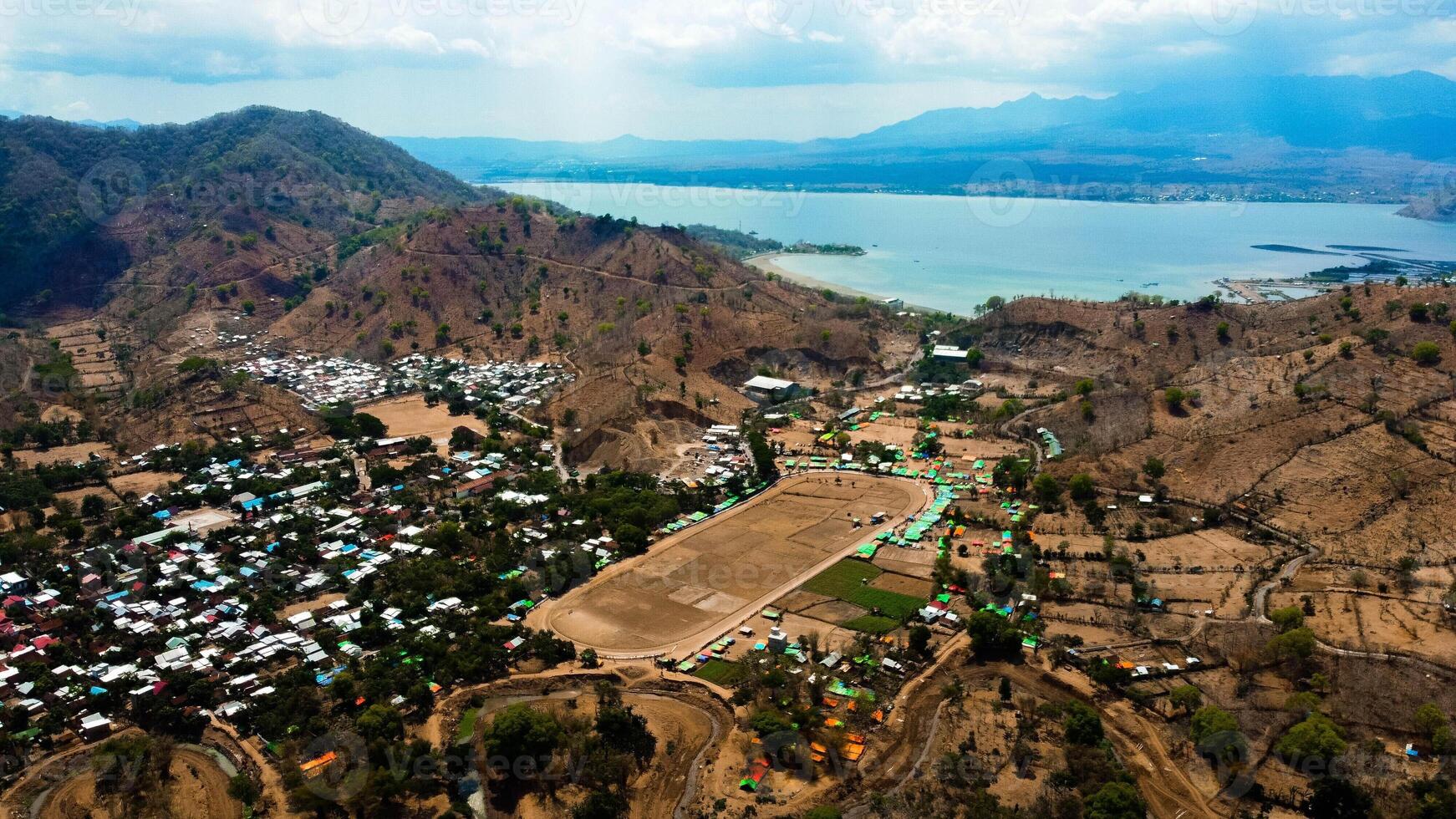 An aerial view of Bima's horse racing field, bustling with majestic horses in competition. The crowd cheers under the bright midday sun, capturing the excitement. photo