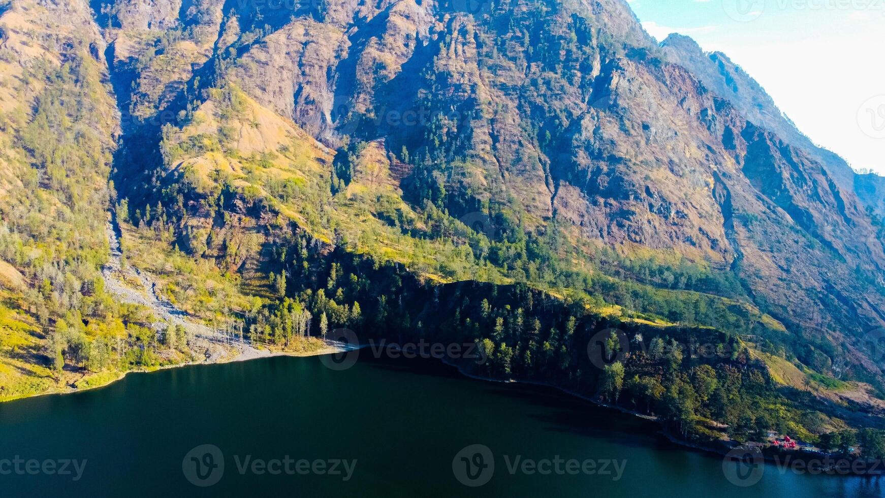 Aerial from the summit of Rinjani shows the stunning Segara Anak Lake below, Surrounded by vibrant blue skies and clouds, the lake's serene beauty stands out against the dramatic landscape. photo